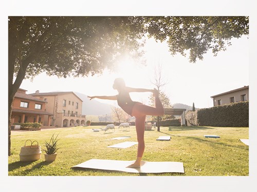 Noia fent exercici de ioga sota un arbre i amb el sol de contrallum, amb el jardi i una masia de fons