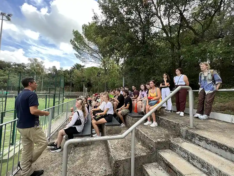 a man giving information to the people seated on the steps of the sports courts