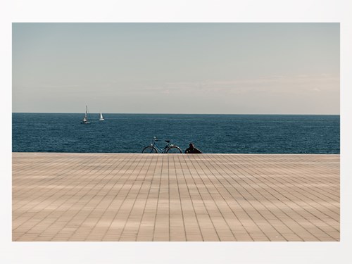 Fotografia d'un noi assegut al costat d'una bicicleta al final d'un passeig marítim i mirant a l'horitzó on passen 2 velers