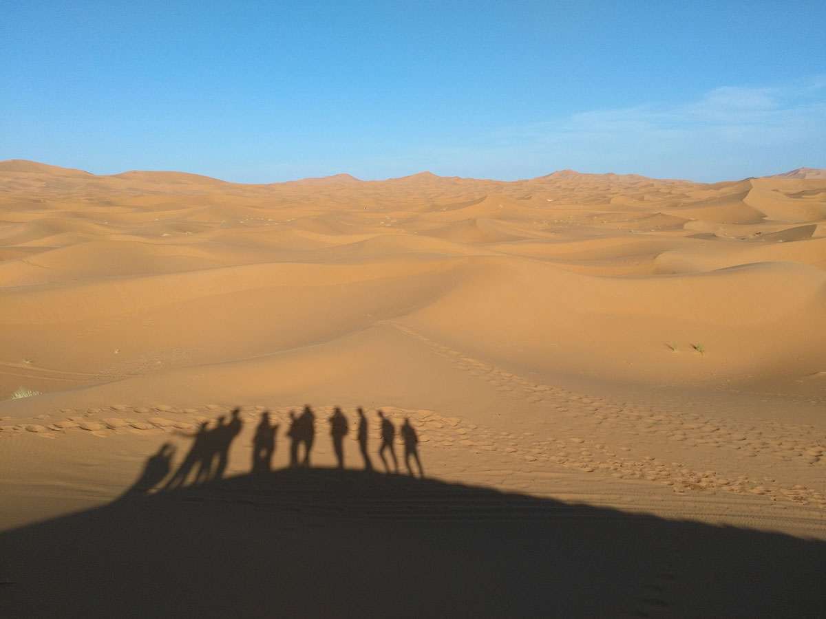 fotografia de paisatge de dunes al desert - a la part inferior es veu l'ombra de les siluetes de la gent del grup projectades a la sorra del desert